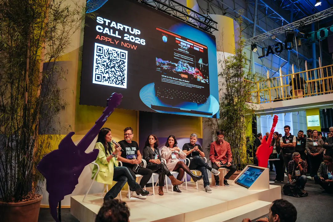 Painel de debate no stand da Smart City of Rock durante a Web Summit, com vários oradores sentados em palco e um ecrã ao fundo a anunciar a “Startup Call 2026 – Apply Now”, acompanhado de um QR code para candidaturas.