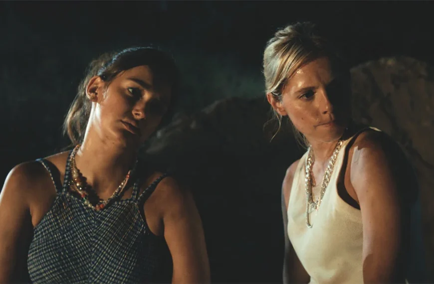 Cena cinematográfica com duas mulheres em conversa intensa, iluminadas por luz quente num ambiente noturno.