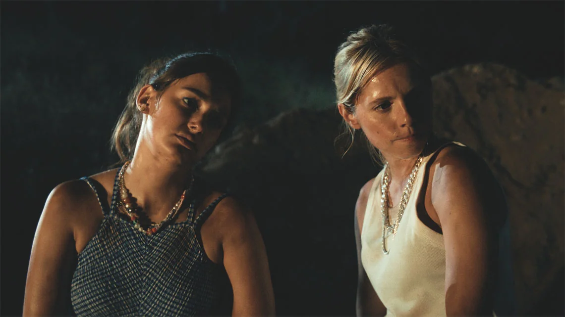 Cena cinematográfica com duas mulheres em conversa intensa, iluminadas por luz quente num ambiente noturno.