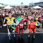 Nicolò Bulega, Yari Montella e Lorenzo Baldassarri celebram no pódio do WorldSBK 2026 em Phillip Island com os troféus e capacetes, perante os fãs.