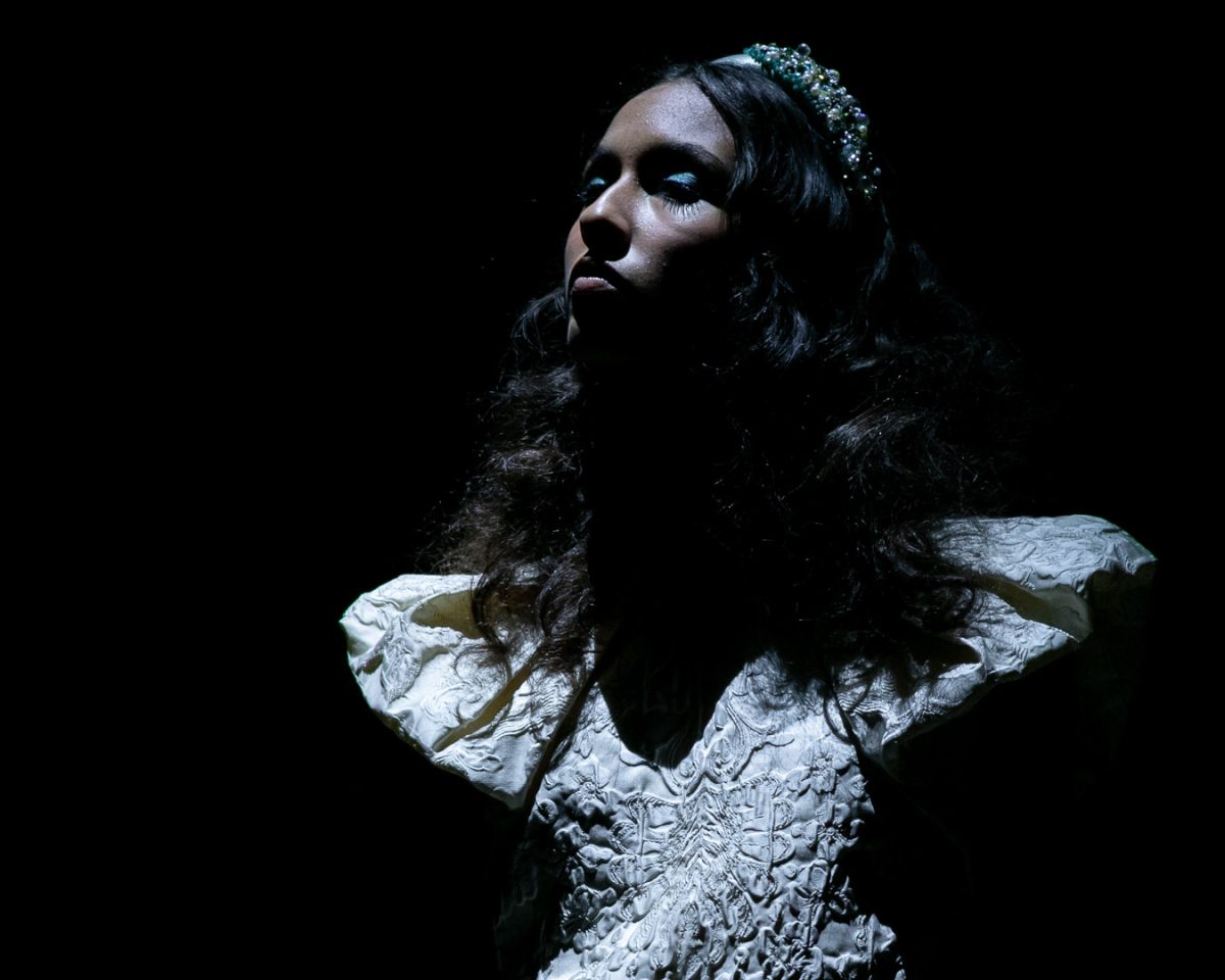 Modelo com vestido de noiva branco e mangas estruturadas desfila sob luz dramática no desfile de Nuno Baltazar na ModaLisboa “Pebbling”.