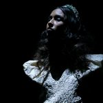 Modelo com vestido de noiva branco e mangas estruturadas desfila sob luz dramática no desfile de Nuno Baltazar na ModaLisboa “Pebbling”.