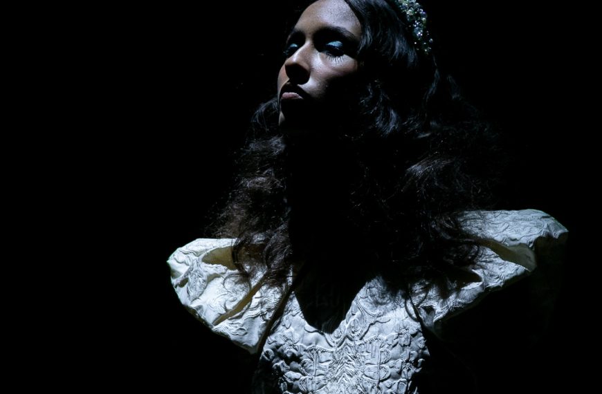 Modelo com vestido de noiva branco e mangas estruturadas desfila sob luz dramática no desfile de Nuno Baltazar na ModaLisboa “Pebbling”.