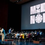 Dino D’Santiago atua com orquestra no espetáculo “Por um Mundo Nôbu” na Gulbenkian, em Lisboa, com o nome do evento projetado em palco.